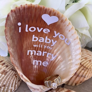 May include: A seashell with the words "I love you baby will you marry me" written on it. A heart shape is above the text. A gold ring with a large diamond is inside the shell.