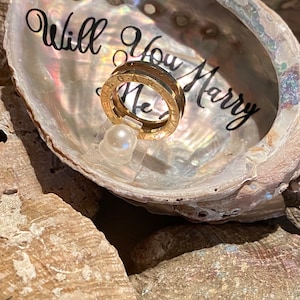 May include: A gold ring with a band of Roman numerals sits inside a white seashell with the words "Will You Marry Me" written inside.