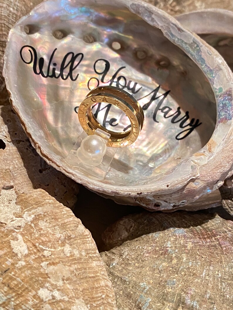 May include: A gold ring with a band of Roman numerals sits inside a white seashell with the words "Will You Marry Me" written inside.