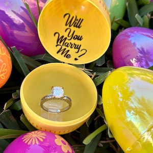 May include: A yellow plastic Easter egg with the words "Will You Marry Me?" written inside. The egg is open and a diamond engagement ring is inside.