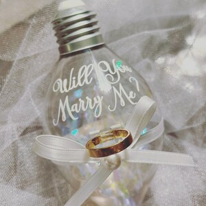 May include: A clear glass light bulb ornament with a white ribbon and a gold ring inside. The bulb has the words "Will You Marry Me?" written on it.
