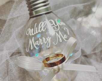Op de afbeelding: Een heldere glazen lichtbollen ornament met een wit lint en een gouden ring erin. De bol heeft de woorden "Will You Marry Me?" erop geschreven.