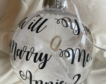 Op de afbeelding: Een transparante glazen kerstbal met witte tule stof erin. De kerstbal heeft een zwarte tekst met de woorden "Will you marry me Annie?"