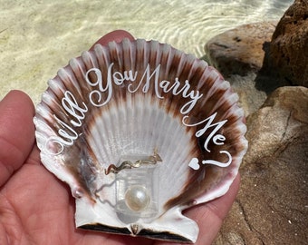 May include: A seashell with the words "Will You Marry Me?" written on it. A ring is inside the shell.
