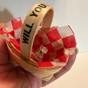 May include: A small wooden basket with a red and white checkered lining. The basket has a handle with the words "WILL YOU" written on it. A ring with a large, clear stone is inside the basket.