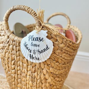 Op de afbeelding: Een geweven strandtas van riet met twee handvatten, gevuld met sandalen. Een wit schelpvormig label is bevestigd met raffia en leest "Please leave Shoes & Sand Here" in zwart schrift. De tas staat op een geweven mat.