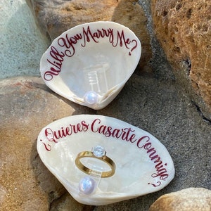 May include: Two white seashells with red lettering. The top shell says "Will You Marry Me?" and the bottom shell says "¿Quieres Casarte Conmigo?"  A gold ring with a diamond is displayed in the bottom shell.