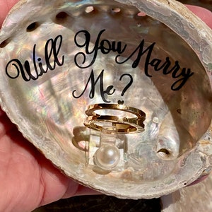 May include: A seashell ring box with the words "Will You Marry Me?" written on the inside. A gold ring and a pearl are inside the box.
