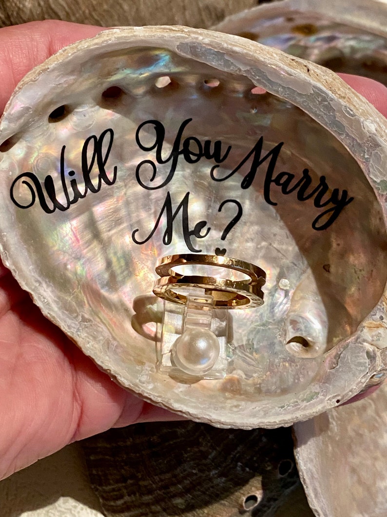 May include: A seashell ring box with the words "Will You Marry Me?" written on the inside. A gold ring and a pearl are inside the box.