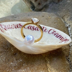 May include: A gold engagement ring with a large, clear gemstone sits in a white seashell. A small, white pearl rests below the ring. The shell has red text that reads "Quieres casarte conmigo."