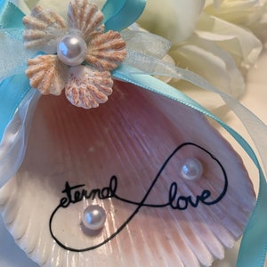 May include: A seashell ring holder with a light pink and white interior. The shell has an infinity symbol drawn on it with the words "eternal love" written inside the symbol. The shell is decorated with a white flower made of seashells and three white pearls.
