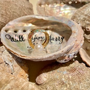 May include: A gold ring with the words "Will You Marry Me" inscribed on it, displayed in a seashell with a pearl.