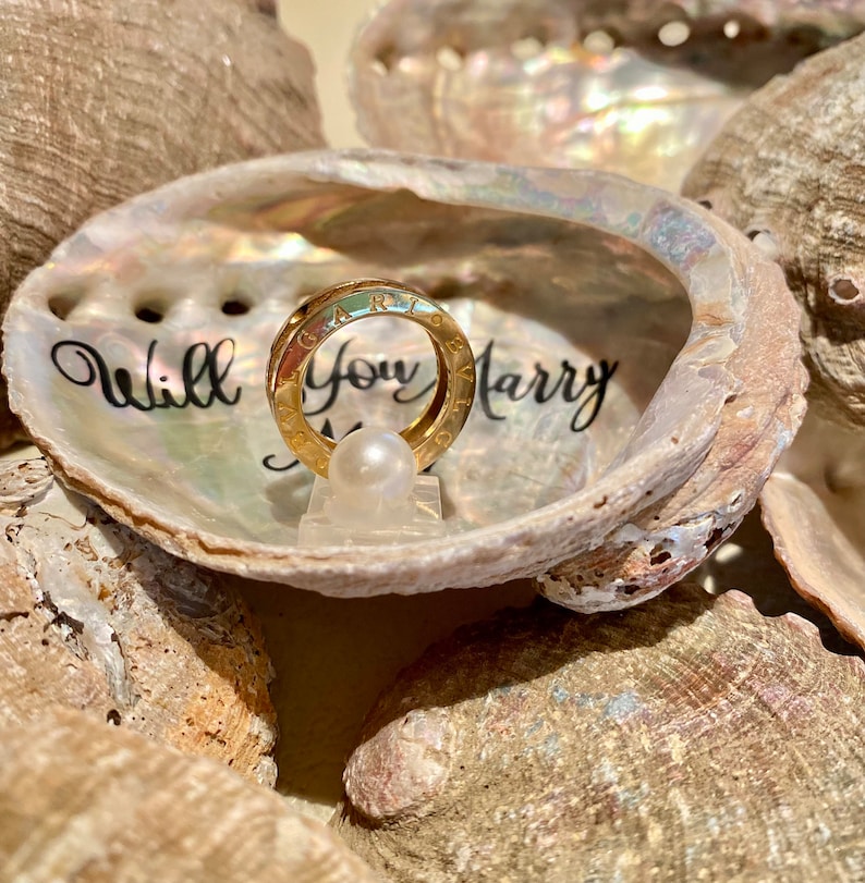 May include: A gold ring with the words "Will You Marry Me" inscribed on it, displayed in a seashell with a pearl.