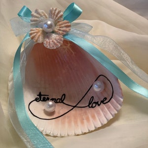 May include: A seashell with a light pink and white color scheme, decorated with a white pearl flower and a ribbon. The shell has the words "eternal love" written on it in black.