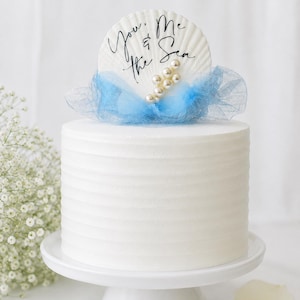 May include: A white cylindrical cake topped with a large white seashell featuring the black script text You & Me & the Sea. The shell is decorated with a cluster of small white pearls and is wrapped in a light blue tulle fabric accent.