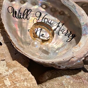 May include: A gold ring with the words "Will You Marry Me" engraved on it, displayed in a white seashell with a pearl.