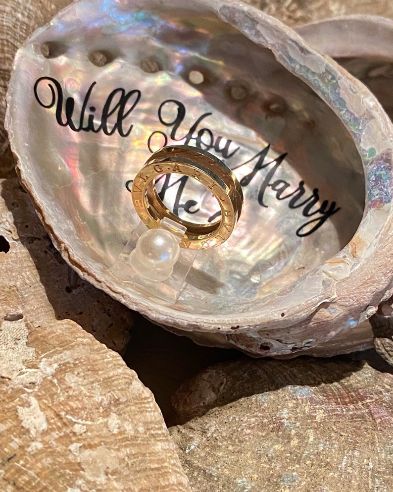 May include: A gold ring with the words "Will You Marry Me" engraved on it, displayed in a white seashell with a pearl.