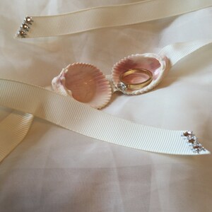 May include: A gold ring with a clear stone rests inside a pink seashell. The seashell is nestled on a white fabric, accompanied by ivory ribbons with rhinestone accents. The ring is a symbol of commitment.