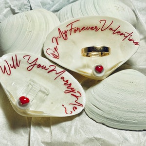 May include: Three white seashells with red writing. The top shell has a gold ring and the words "Be My Forever Valentine". The middle shell has the words "Will You Marry Me?" and the bottom shell is blank.