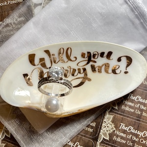 May include: A seashell ring dish with the words "Will you marry me?" written in gold lettering. A diamond engagement ring is displayed on the dish with a pearl next to it.