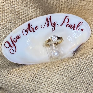 May include: A white seashell ring dish with the words "You Are My Pearl" written in red cursive. A gold ring with a clear stone is displayed on the dish, next to a white pearl.