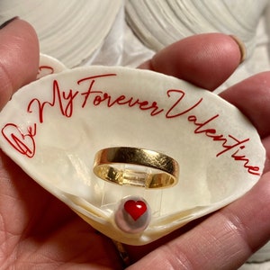 May include: A white seashell with the words "Be My Forever Valentine" written in red. A gold ring and a pearl with a red heart are inside the shell.