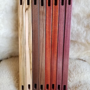 Puede incluir: Una colección de ocho asas de madera para bolsos en varios colores, incluyendo marrón claro, marrón, marrón rojizo y morado. Cada asa tiene forma rectangular con una ranura en cada extremo. Las asas se muestran sobre una superficie blanca texturizada.