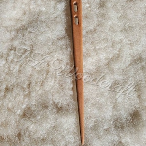 May include: A wooden needle with two holes near the top, designed for spinning yarn. The needle is brown and has a pointed tip. It is lying on a white fluffy surface.