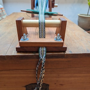 Peut inclure: Un métier à tisser en bois avec une bande partiellement tissée à motifs verts, blancs et marron. Le métier à tisser a un cadre en bois, un rouleau en bois et du matériel métallique. La bande tissée pend du métier à tisser. Le métier à tisser est posé sur une surface en bois.