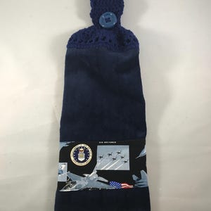 May include: Navy blue kitchen towel with a crocheted top and button closure. The towel features a decorative band with U.S. Air Force imagery, including jets, the Air Force seal, and an American flag design.