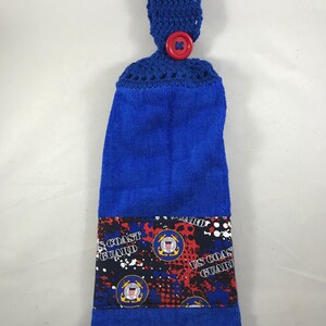 May include: Blue kitchen towel with a crocheted hanging loop and a red button. The towel has a printed panel with the US Coast Guard emblem and the words "US Coast Guard" repeated.
