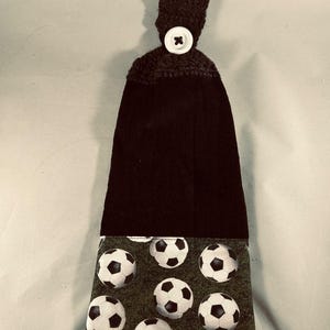 May include: A black kitchen towel with a soccer ball pattern at the bottom. The top portion is crocheted with a white button closure. The soccer ball pattern features white and black soccer balls on a green background.
