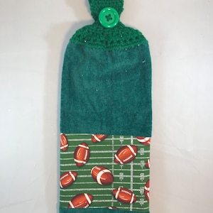 May include: Green kitchen towel with a green crocheted hanging loop and a green button. The towel has a green and white football field print with brown footballs.