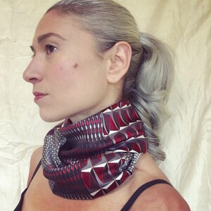 Könnte beinhalten: Eine Frau trägt einen roten, schwarzen und weißen gemusterten Halstuch. Das Tuch ist aus seidenartigem Material und hat ein geometrisches Muster.