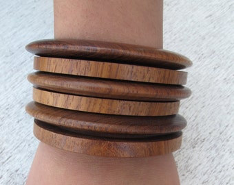 May include: A stack of five wooden bangles in a warm brown colour. The bangles are stacked on a person's wrist.