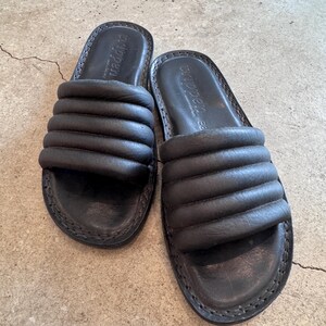 May include: A pair of black leather slide sandals. The sandals have a padded, horizontal-striped design across the top. The soles are stitched with a visible seam. The brand name "crippen" is embossed on the footbed.