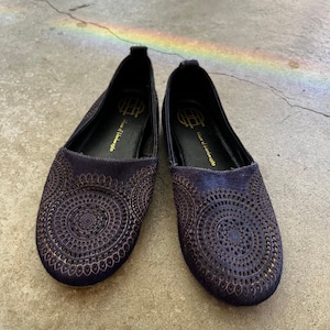 May include: A pair of dark purple leather flats with intricate circular cutouts and gold detailing. The shoes have the text "House of Leather 1960" on the inside.