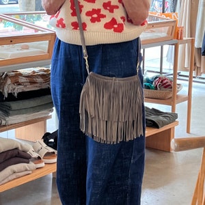 May include: A gray fringed suede crossbody bag with a gold chain strap. The bag is worn over a white sweater with red floral print and blue denim pants.