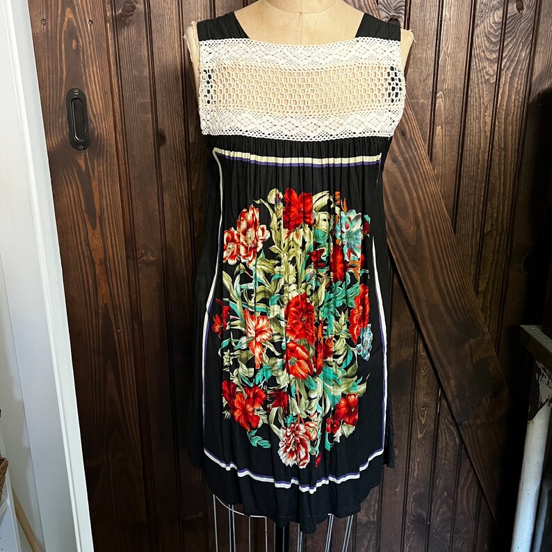 Pu&ograve; includere: Abito nero con corpetto in crochet bianco e stampa floreale. L'abito presenta uno scollo tondo e una silhouette aderente. La stampa floreale comprende fiori rossi, verdi e blu. Le finiture bianche corrono lungo i lati dell'abito.