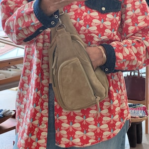 May include: A tan suede crossbody bag with a front pocket and zipper closure. The bag is worn over a red button-up shirt with a white and blue butterfly pattern and denim accents. The person is also wearing blue jeans.