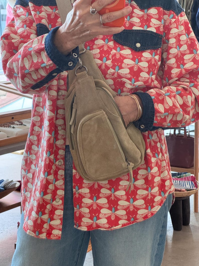 May include: A tan suede crossbody bag with a front pocket and zipper closure. The bag is worn over a red button-up shirt with a white and blue butterfly pattern and denim accents. The person is also wearing blue jeans.