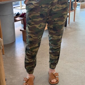 May include: Camouflage print trousers with an elasticated waistband and cuffs. The trousers are worn with brown leather sandals with a silver buckle. The outfit has a casual and relaxed style.