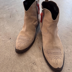 May include: Beige suede ankle boots with decorative stitching on the toe and a side zip with a red pull. The boots have a pointed toe and a low heel. Made in Italy.