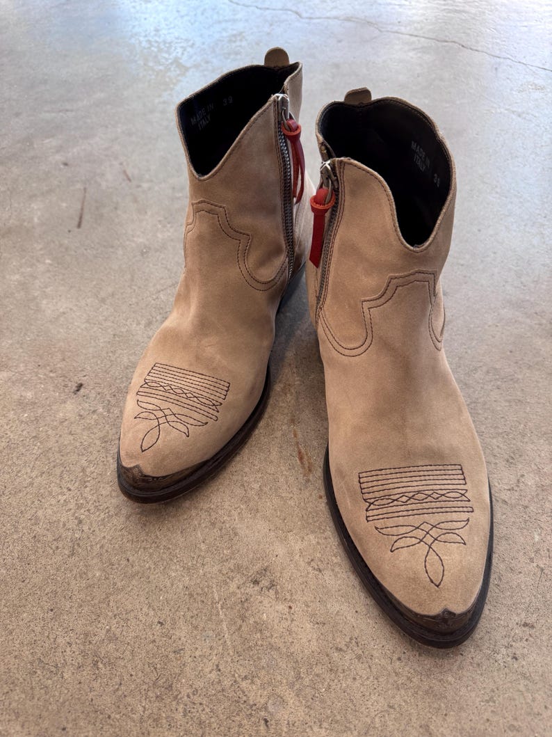 May include: Beige suede ankle boots with decorative stitching on the toe and a side zip with a red pull. The boots have a pointed toe and a low heel. Made in Italy.