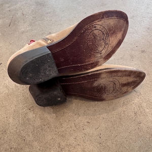 May include: A pair of beige suede ankle boots with dark brown soles. The soles feature a circular logo with text. The boots have a small heel and a zipper detail. The boots are on a concrete surface.