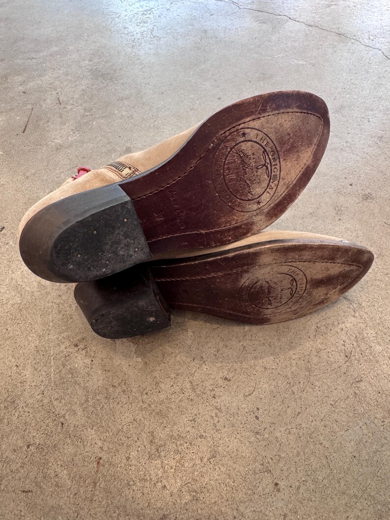 May include: A pair of beige suede ankle boots with dark brown soles. The soles feature a circular logo with text. The boots have a small heel and a zipper detail. The boots are on a concrete surface.