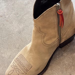 May include: A tan suede ankle boot with a pointed toe and decorative stitching. The boot has a side zip with a red leather pull tab. The words "MADE IN ITALY" are visible inside the boot.