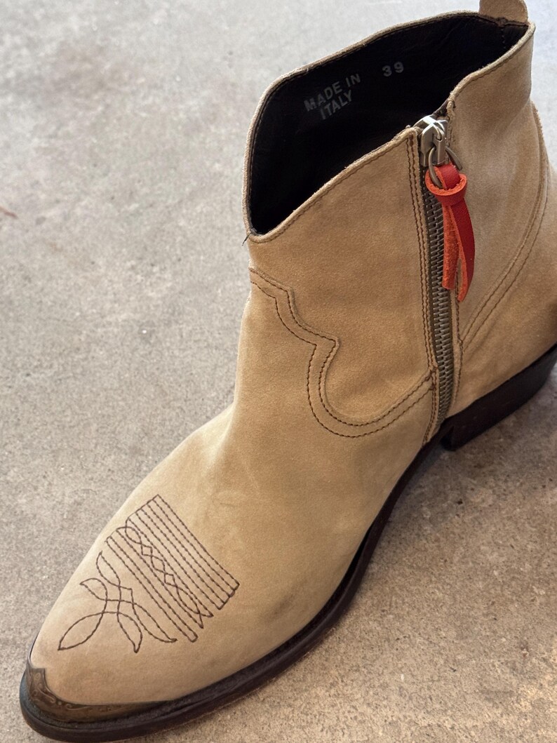 May include: A tan suede ankle boot with a pointed toe and decorative stitching. The boot has a side zip with a red leather pull tab. The words "MADE IN ITALY" are visible inside the boot.