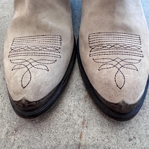 May include: A pair of light tan suede cowboy boots. The boots feature decorative stitching on the toe and vamp, with a dark brown leather sole and metal toe caps. The boots are a neutral colour.