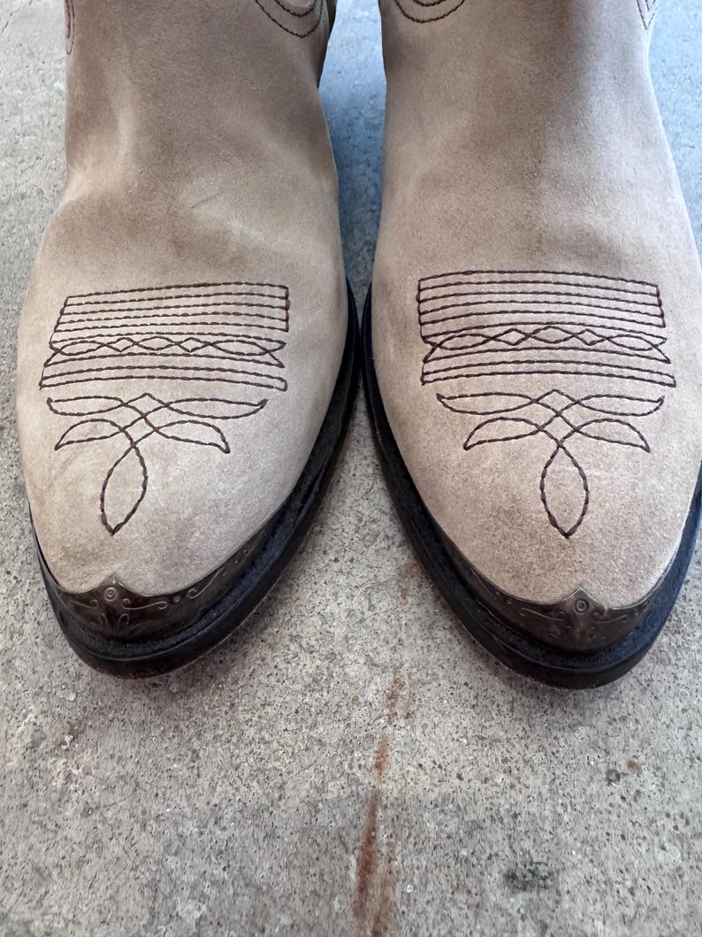 May include: A pair of light tan suede cowboy boots. The boots feature decorative stitching on the toe and vamp, with a dark brown leather sole and metal toe caps. The boots are a neutral colour.